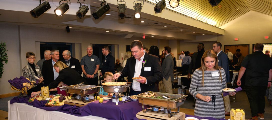 Guests serving themselves food from a buffet table during a catered event at the Knight Club at Alfred University, under stage lighting in a modern event space.
