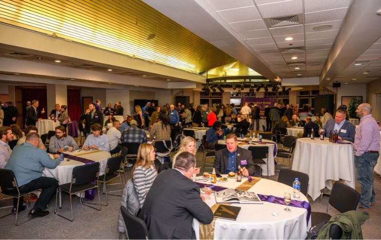 Crowd of guests socializing and dining at round tables during an event inside the Knight Club at Alfred University, with stage lighting and a high ceiling overhead.