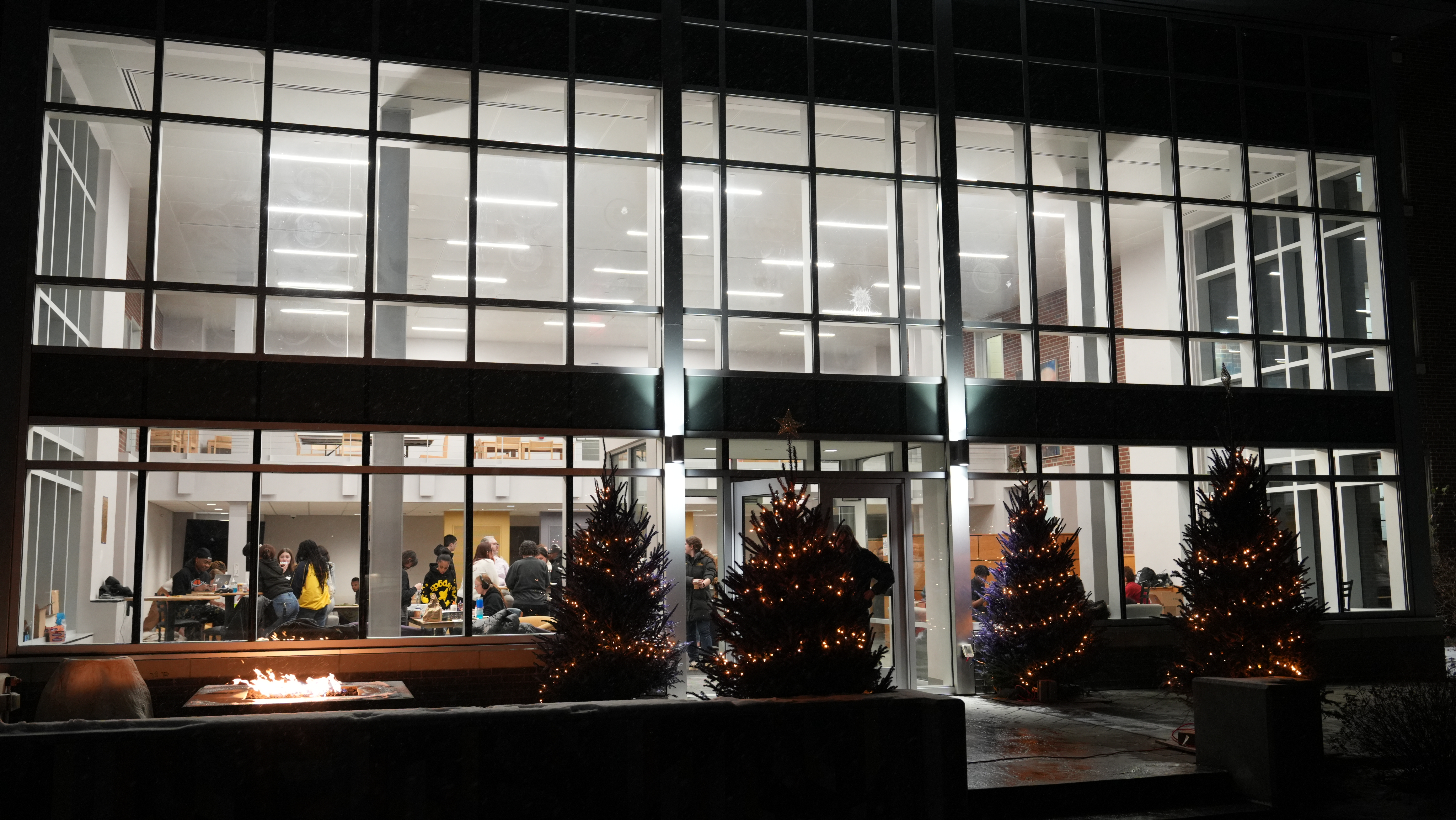 The Link residence hall at night, with holiday lights on trees outside and students gathered inside around tables