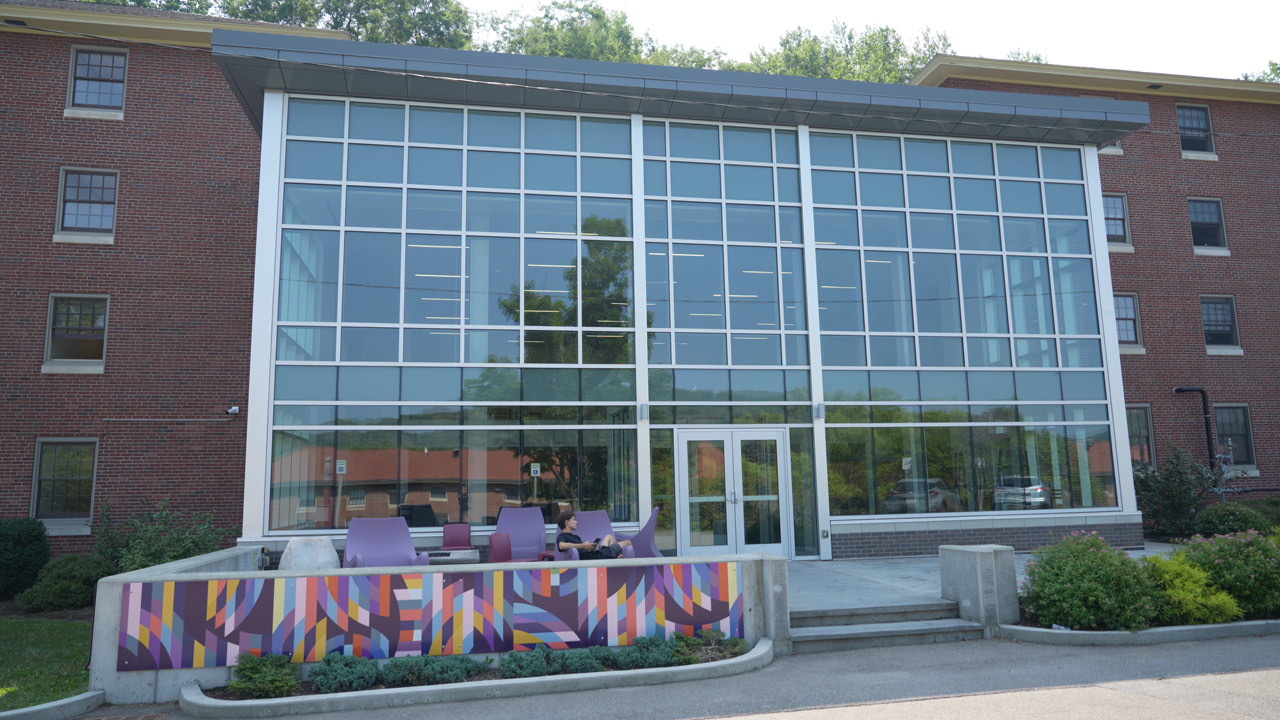 Front view of The Link residence hall with large glass windows, modern outdoor seating, and colorful mural panels at Alfred University.