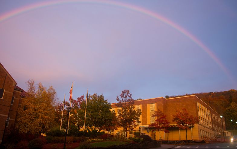 McMahon Engineering Building at Alfred University illuminated by sunset light, with trees in fall colors and a full rainbow arching across the sky above the campus