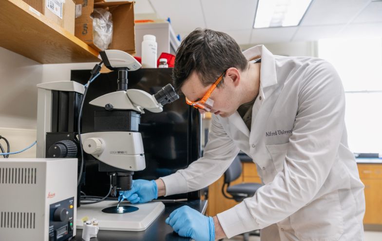 Alfred University student in a lab coat and safety goggles using a microscope in a research lab inside the McMahon Engineering Building