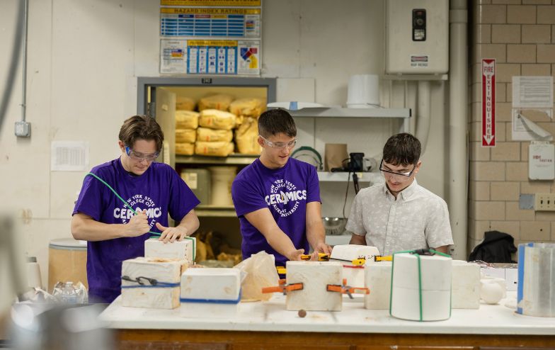 Three Alfred University students working with ceramic molds in a lab space, wearing safety goggles and purple 'Ceramics' shirts, inside the McMahon Engineering Building.