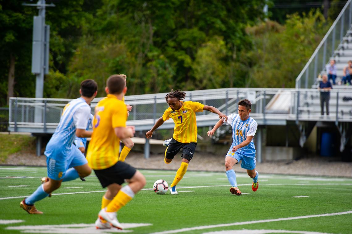 Soccer player in a yellow jersey with number 7 dribbles the ball while being chased by an opponent in a light blue and white striped jersey. Two other players and spectators on bleachers are visible in the background, with trees surrounding the green field.