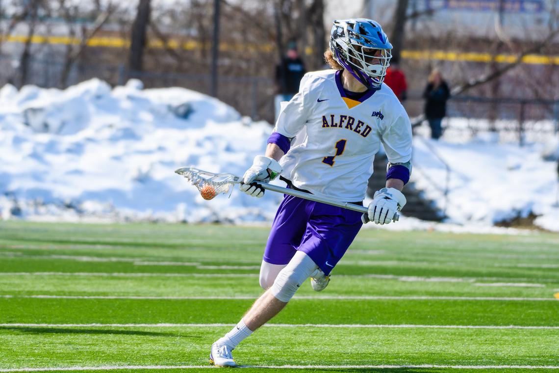 Lacrosse player in a white 'ALFRED' jersey with number 1, purple shorts, and a blue helmet runs on a green field holding a lacrosse stick. Snow, trees, and blurred figures are visible in the background.