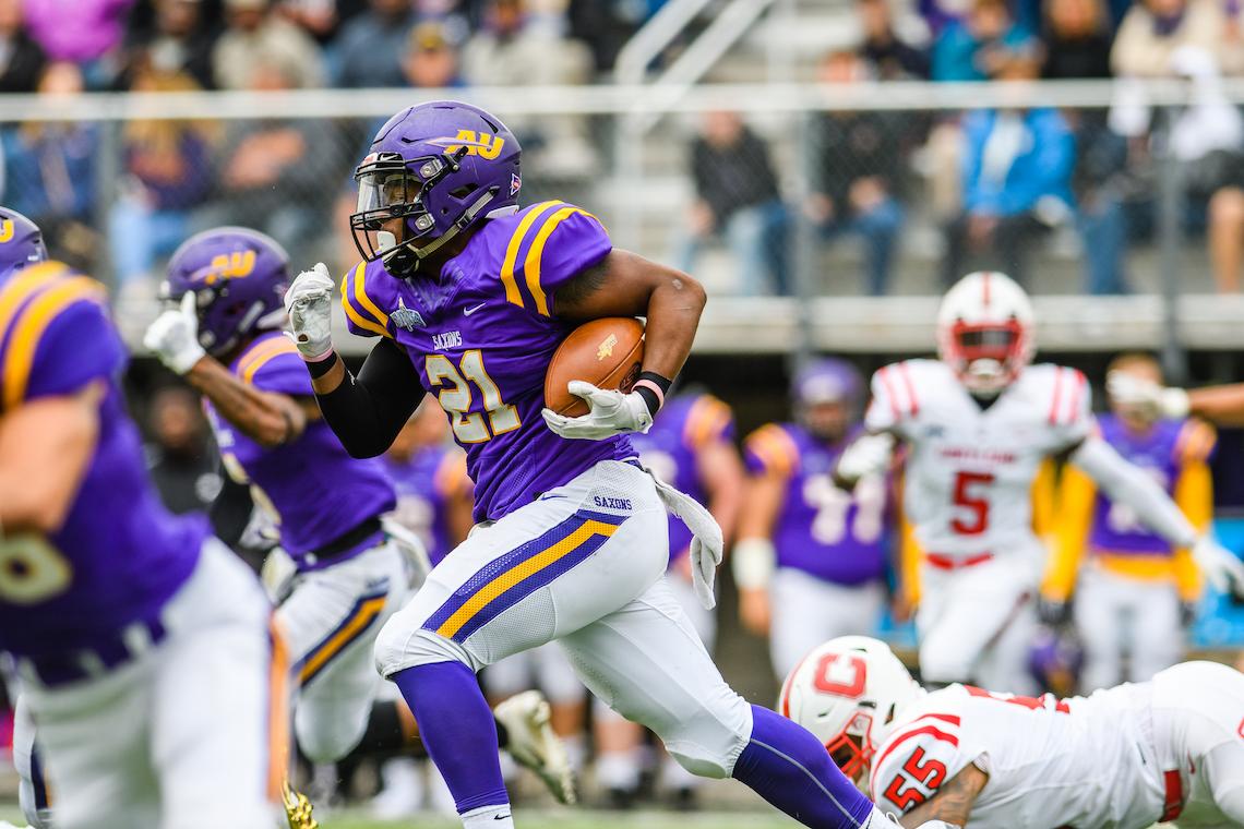 Football player in a purple and yellow uniform, number 21, runs with the ball during a game. A player in a white and red uniform is on the ground reaching toward him, while other players and a crowd of spectators are visible in the background.