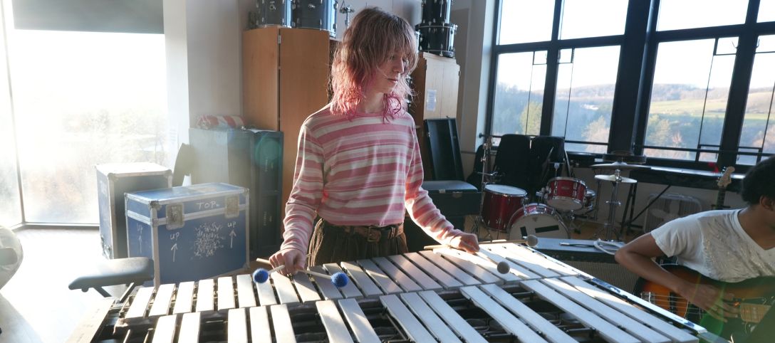Student playing a vibraphone in a sunlit music rehearsal room at Alfred University's Miller Performing Arts Center, with drums and instruments in the background and scenic views through large windows.
