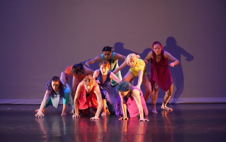 Group of student dancers performing on stage under colorful lighting at Alfred University's Miller Performing Arts Center, wearing vibrant costumes in a choreographed formation.