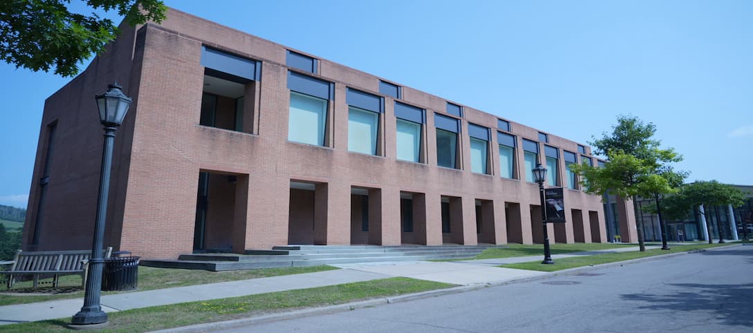 Brick building with upper windows and ground-floor archways, next to a sidewalk with trees, lampposts, and a bench under a clear blue sky.
