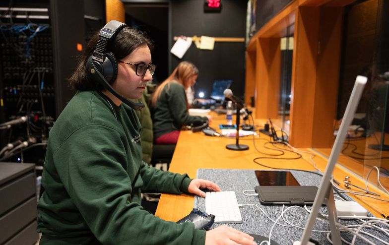Students operating sound and lighting equipment in a control booth at Alfred University's Miller Performing Arts Center, wearing headsets and monitoring a live production.