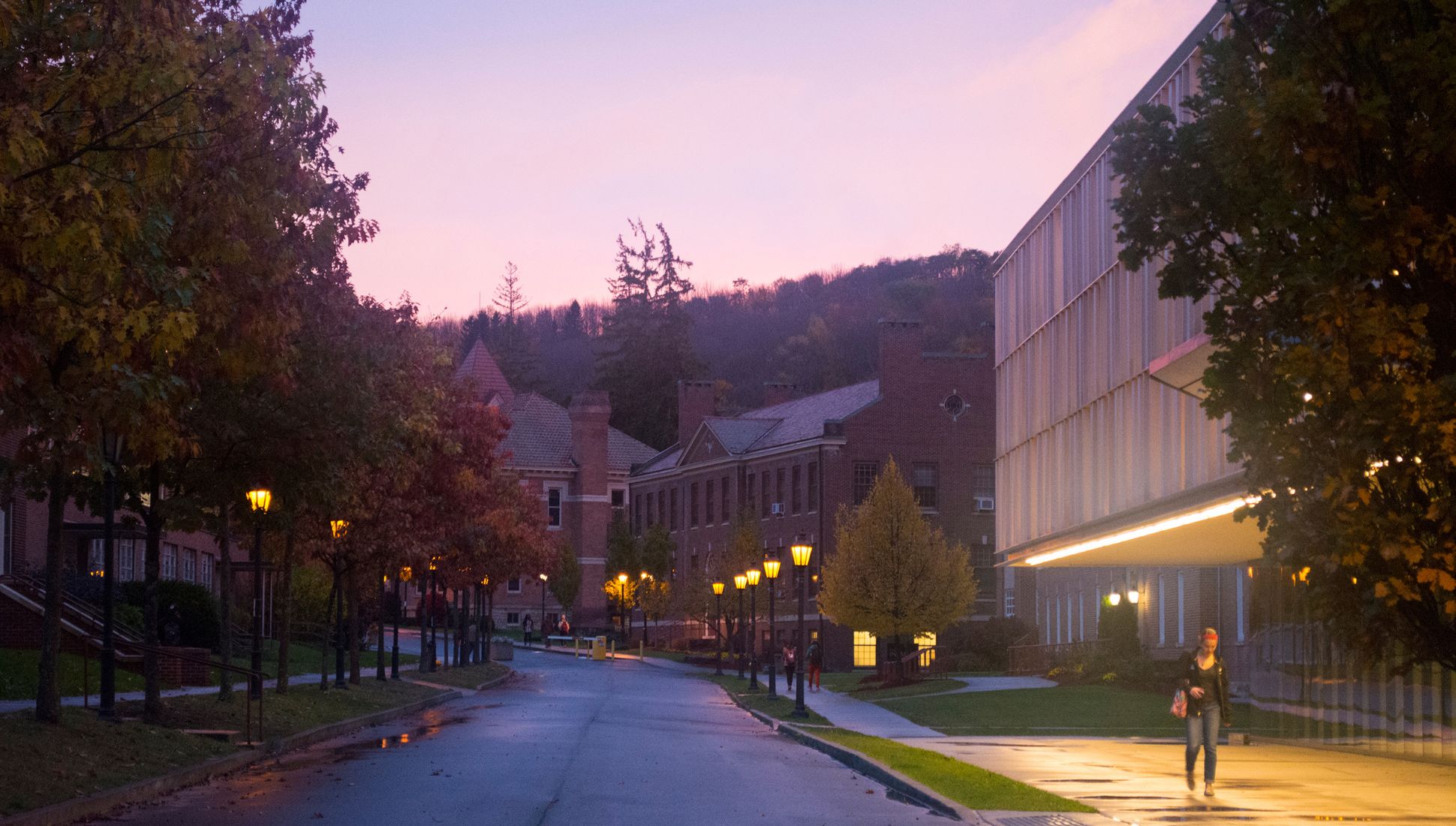 Evening view of Alfred University campus with historic brick buildings, a modern glass-paneled pavilion, and students walking along a tree-lined street under glowing streetlights.