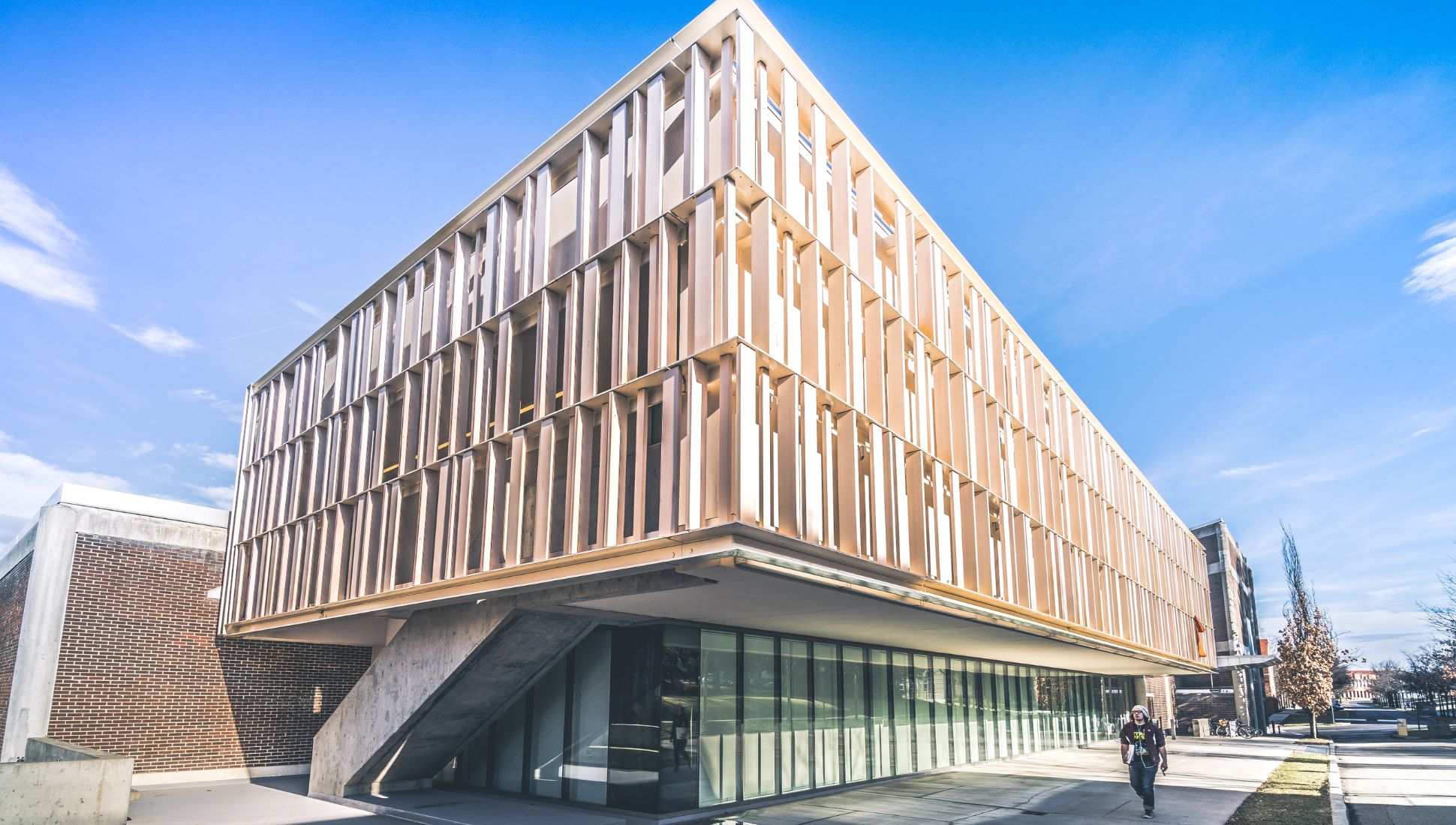 Exterior view of Alfred University’s modern pavilion with vertical slatted facade, angled concrete support, and glass walls, shown under a bright blue sky.