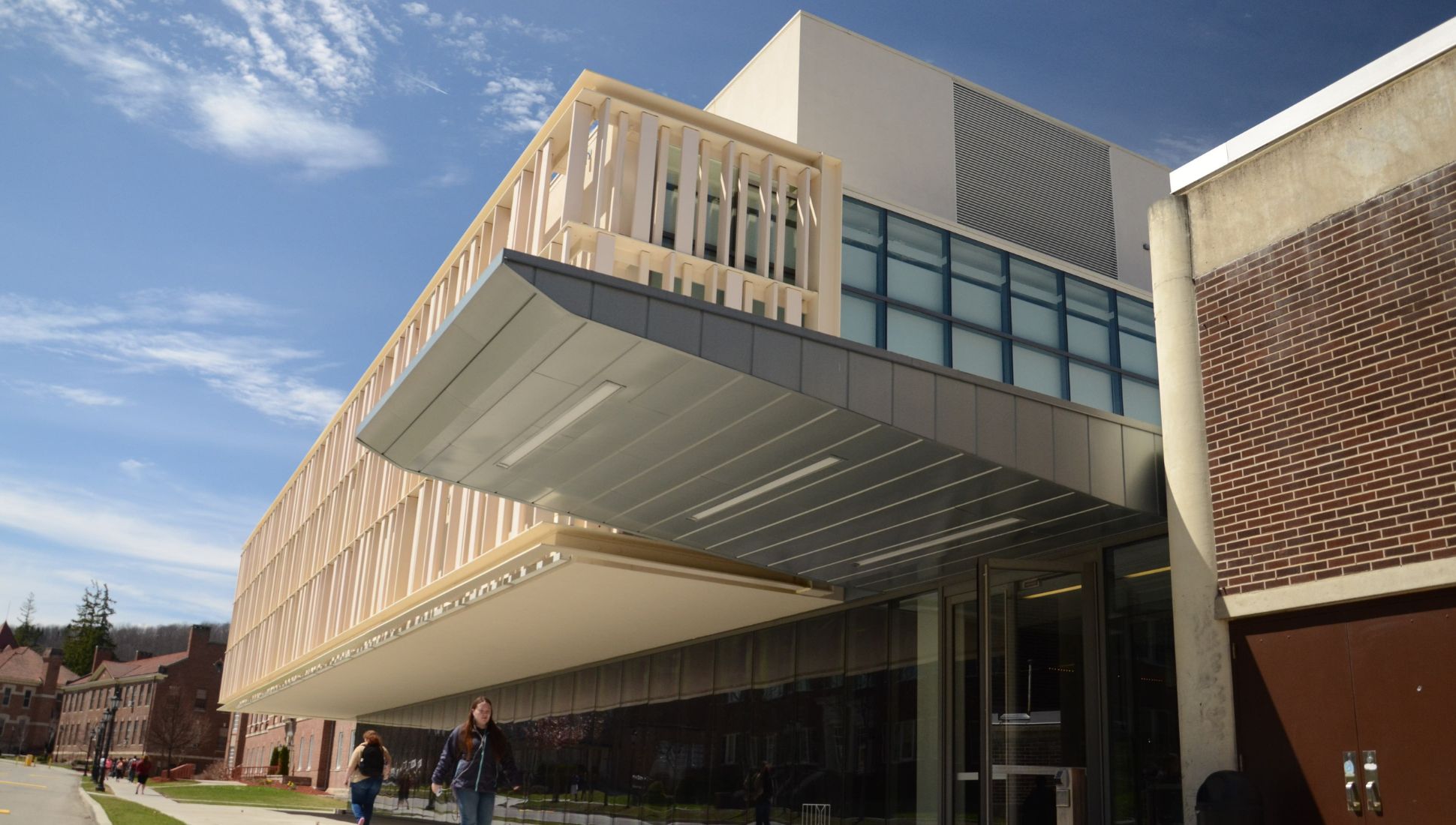 Front view of Alfred University’s modern glass and metal pavilion with cantilevered upper floor, as students walk along the adjacent sidewalk on a sunny day.