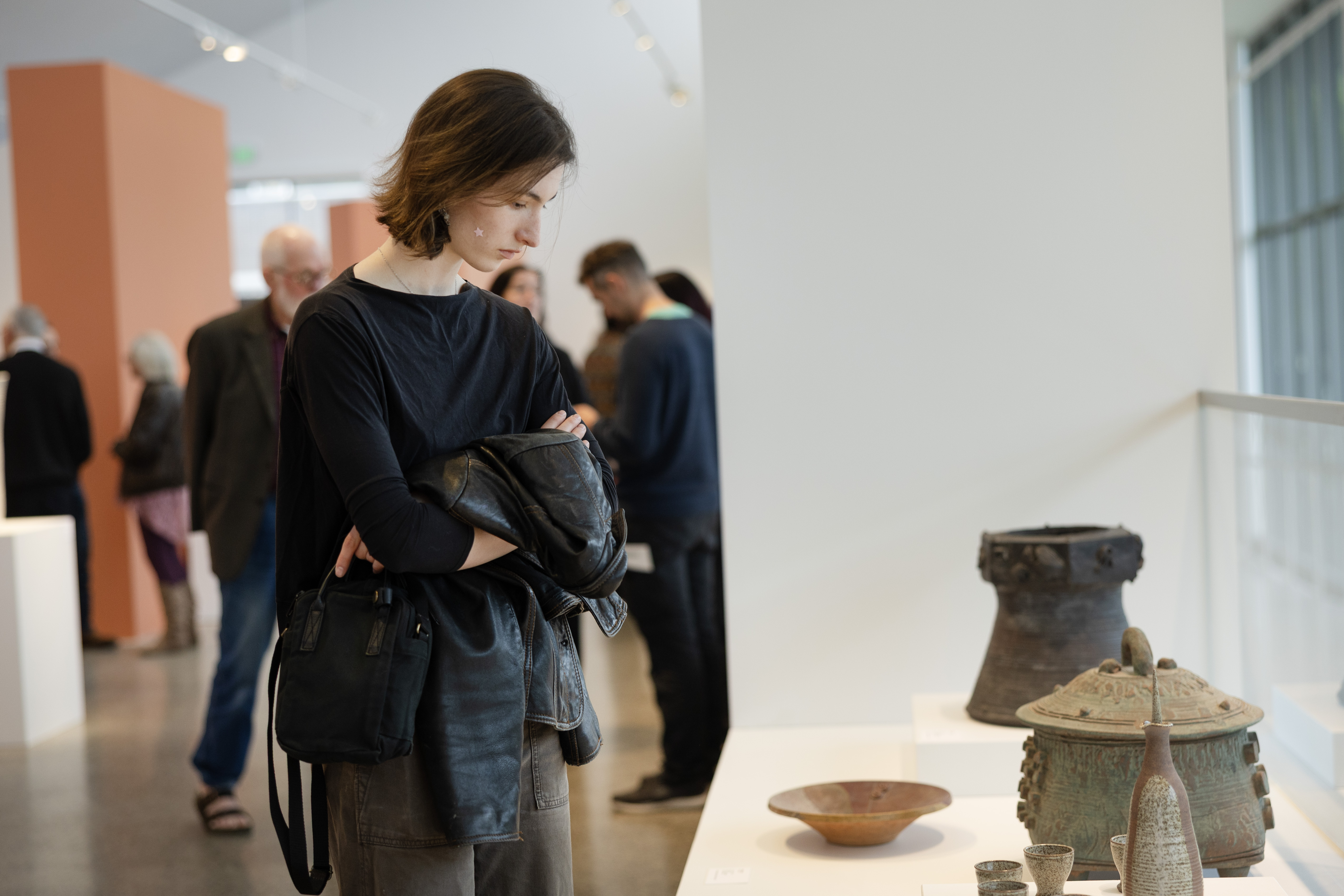 A visitor closely observing ceramic and metal vessels on display in an Alfred University art gallery, with other guests browsing the exhibition in the background.