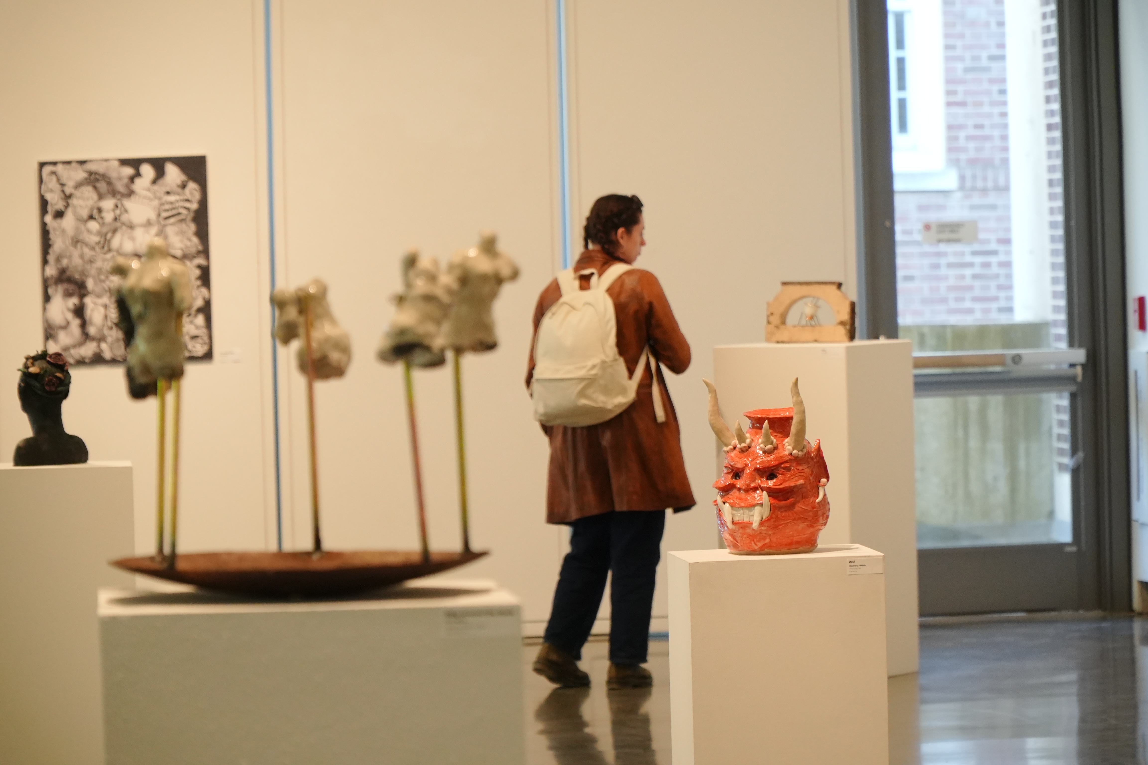 A visitor with a backpack viewing ceramic and mixed-media sculptures in an Alfred University art gallery exhibition.