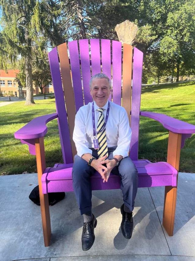 Alfred University President Mark Zupan sits on an oversized purple and brown wooden chair placed on a concrete surface outside of Alumni Hall. He is dressed in a white shirt, navy blue pants, black shoes, and has on a yellow and black striped tie. The background includes green grass, trees, and part of Powell Campus Center on the left. 