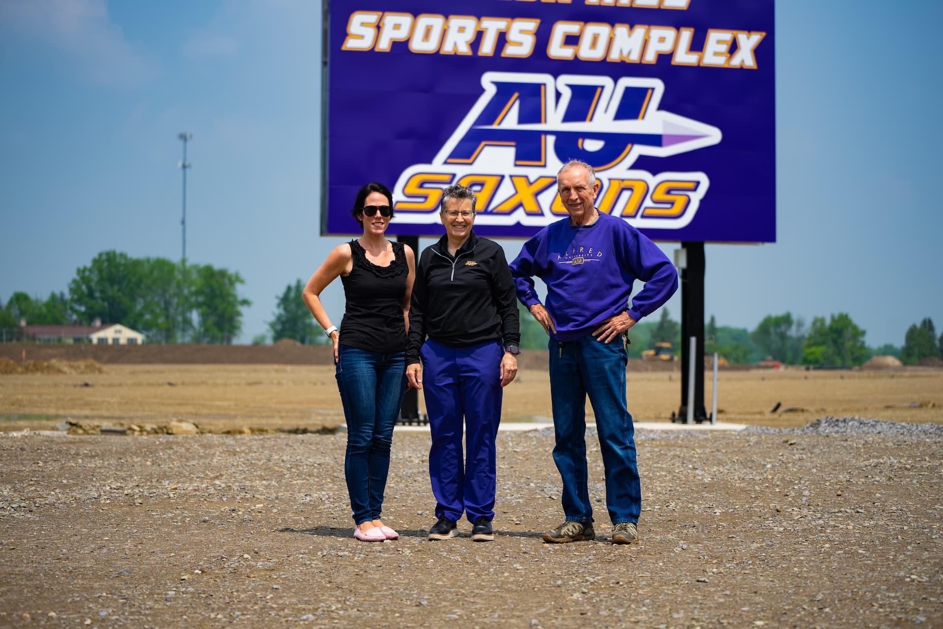 Three people stand on a gravel area in front of a large sign that reads 'SAXON HILL SPORTS COMPLEX' at the top, with 'AU SAXONS' and a logo in the center. Behind them is an open field with trees and buildings in the distance under a clear blue sky.