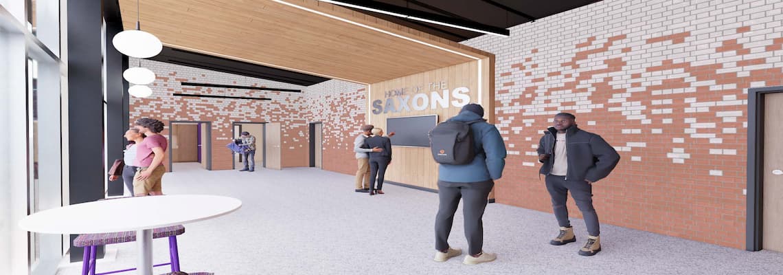 A modern indoor hallway with a high ceiling and large windows on the left side, allowing natural light to fill the space. The walls feature a mix of white and red bricks in a patterned design. Several people are standing and talking in the hallway. On the right wall, a sign reads "HOME OF THE SAXONS" above a flat-screen TV. Multiple doors are visible along the hallway.