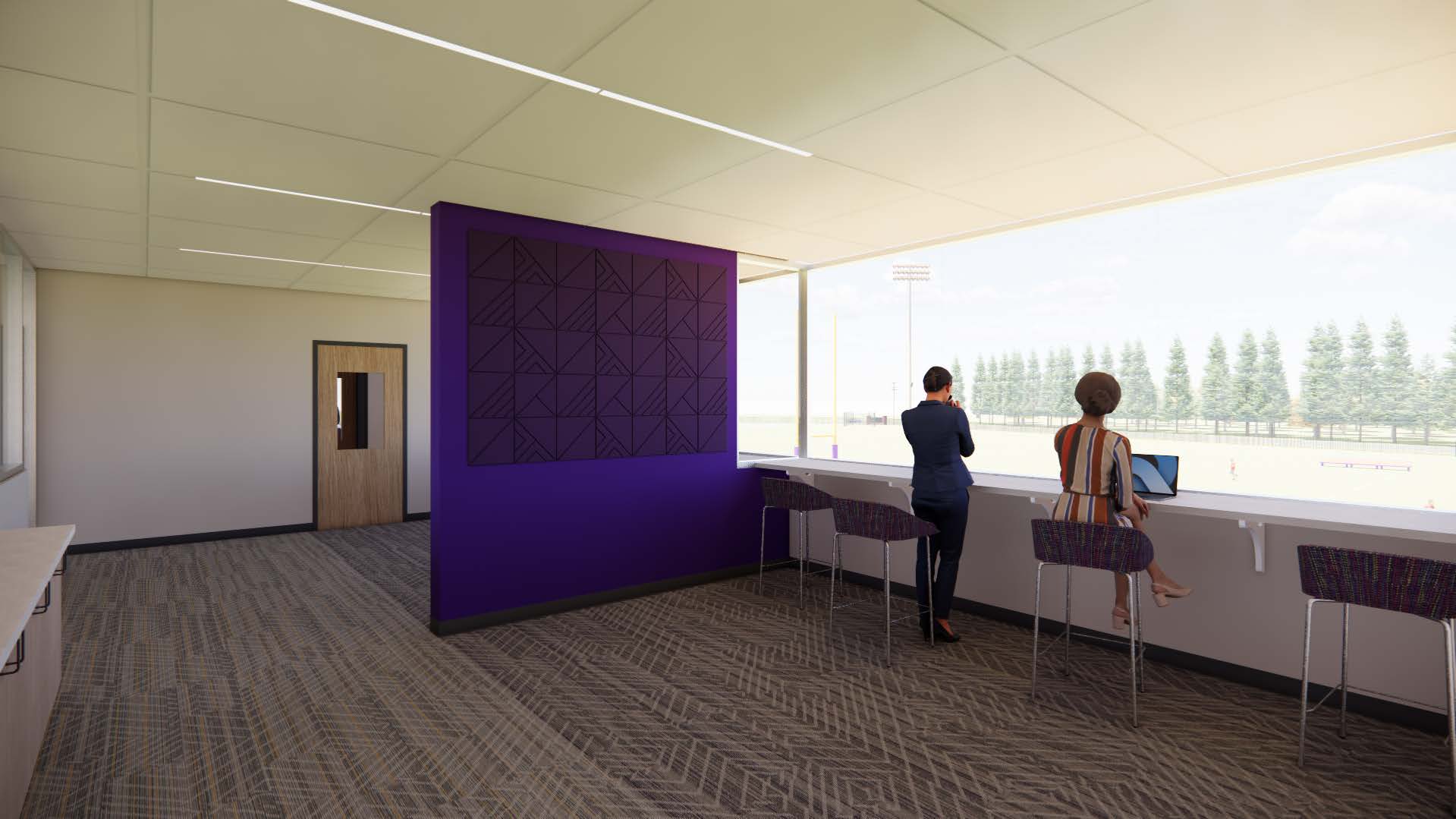 A modern indoor viewing area with large windows overlooking an outdoor sports field. The room features patterned carpeting, a purple accent wall with geometric designs, and several high chairs lined up along a counter facing the window. One person is standing and looking out, while another is seated using a laptop. A door is visible on the left side of the room. 