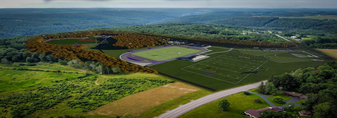 Aerial view of a large sports complex nestled among green hills and dense forests. The complex includes a football field with bleachers, a track and field area, and several practice fields. Surrounding the complex are parking lots, access roads, and a winding road in the foreground leading to nearby buildings. The landscape features a mix of open grassy areas and thick woodland.