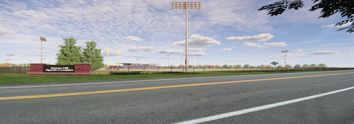 A wide view of an outdoor sports complex taken from across a road. The sports complex is surrounded by a black metal fence and features multiple tall light poles, goalposts, and bleachers in the background. A red brick sign with white text on the left side of the image reads "Saxons Hill Sports Complex." The sky is mostly clear with scattered clouds, and there are trees visible behind the fence.