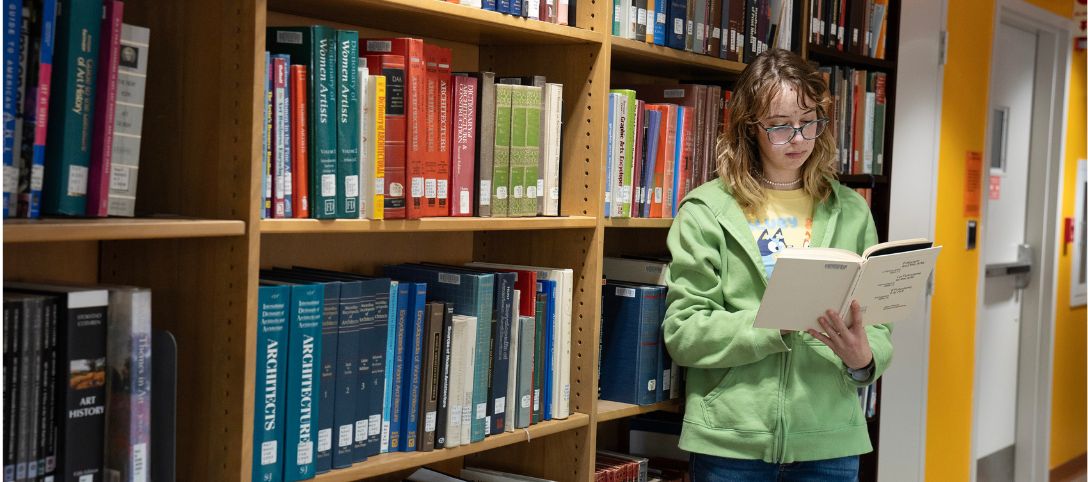A student wearing a green hoodie reads a book while standing in front of bookshelves filled with art, architecture, and design books in Scholes Library.