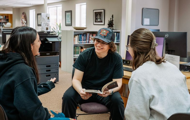 Three students sit in a group at Scholes Library, smiling and talking while one of them holds an open book, with computers and bookshelves in the background.