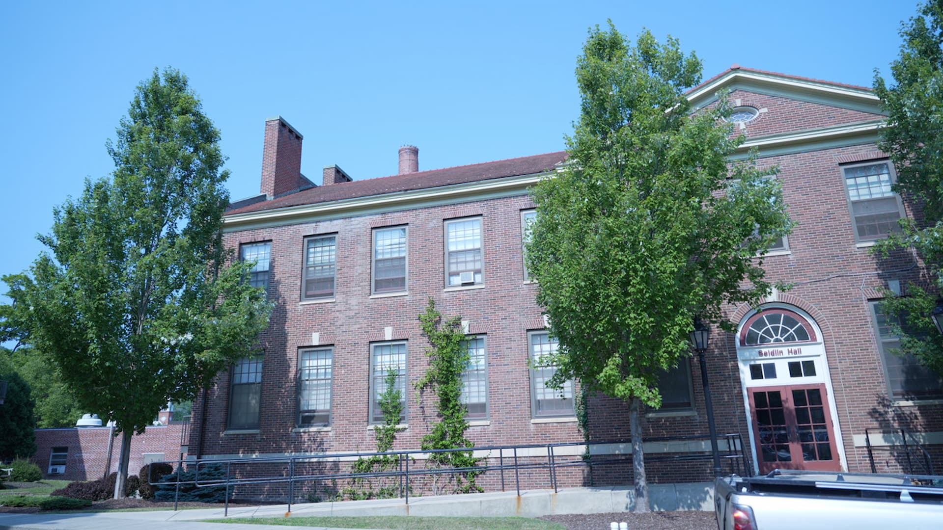 A red-brick building named 'Seldin Hall' with a red roof and several evenly spaced windows. A ramp leads to the main entrance, which features a white-framed glass door beneath a sign that reads 'Seldin Hall.' Leafy trees partially obscure the view, and a vehicle is parked in the lower right corner. The setting suggests a campus environment.