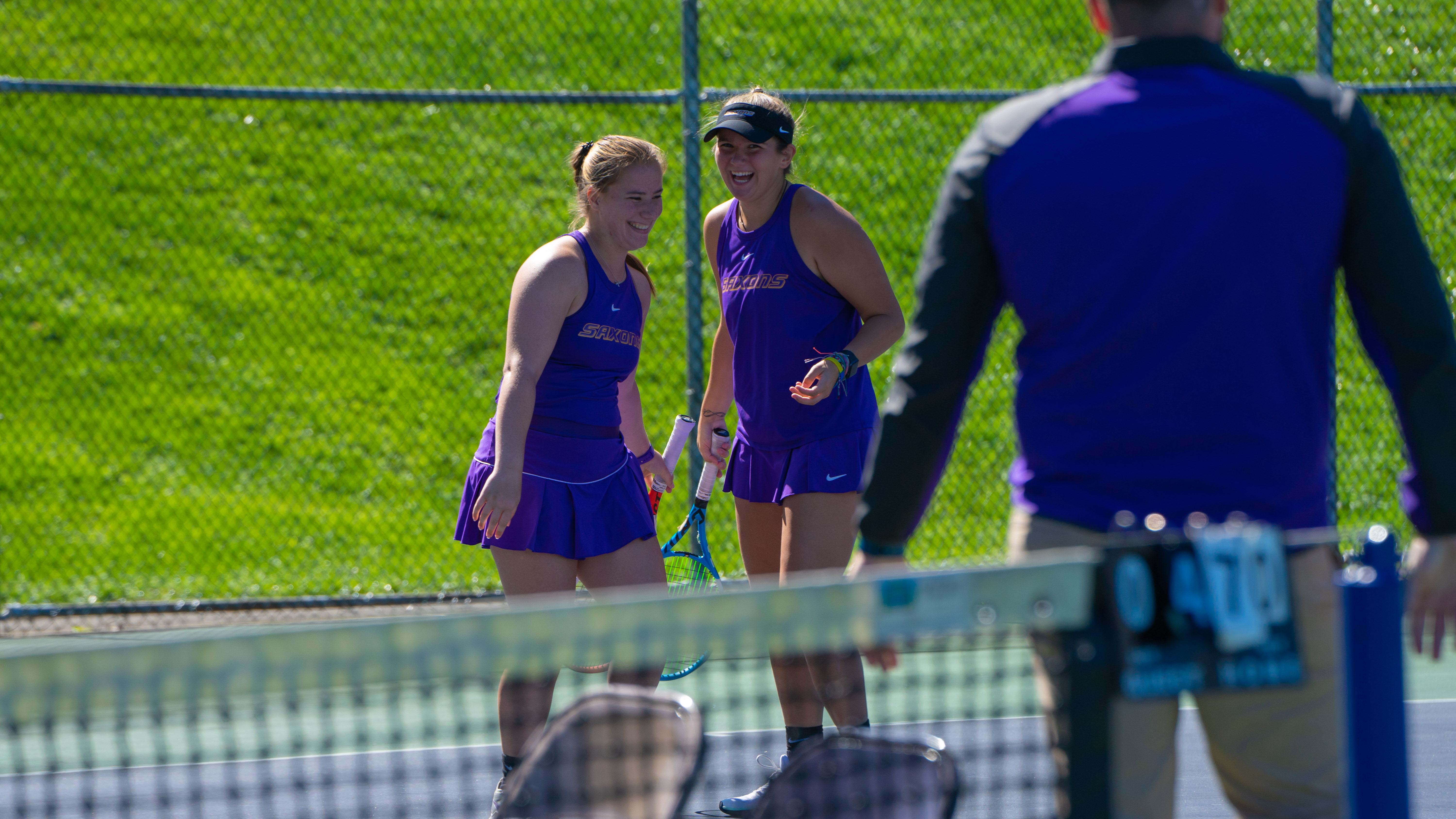 Two female tennis players in purple uniforms smiling and talking during a doubles match, with a coach standing nearby