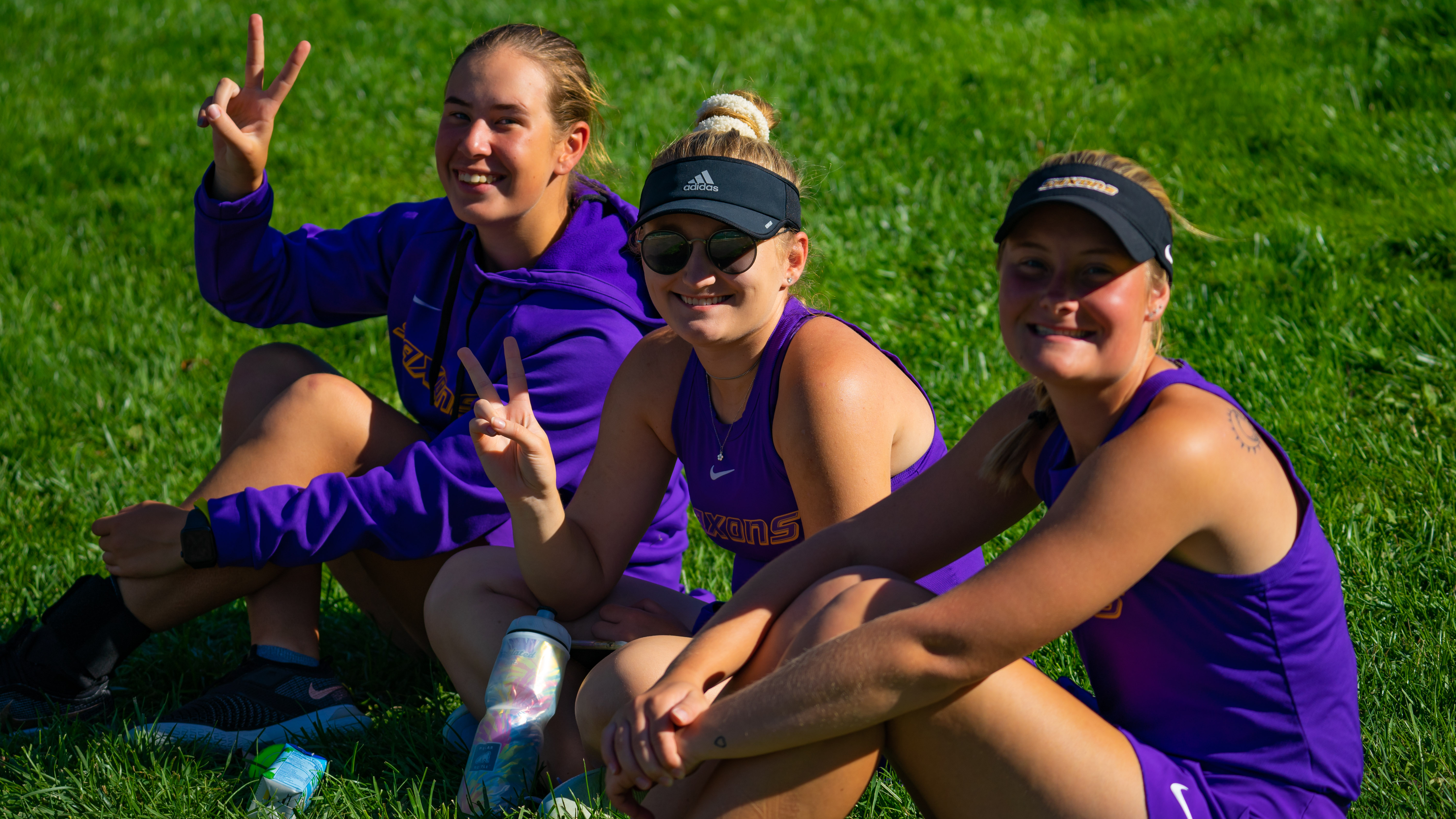 Three female tennis players sitting on the grass, smiling and flashing peace signs while wearing purple Alfred University gear.