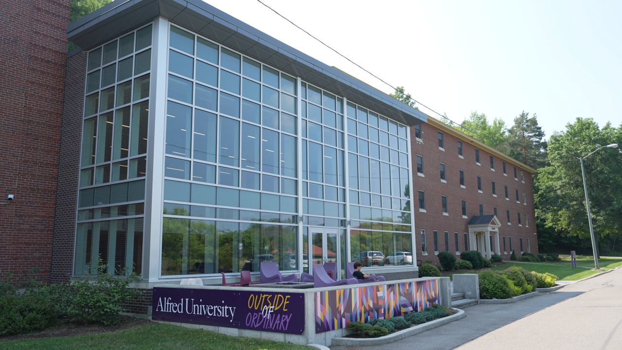 Exterior view of The Link at Alfred University, featuring a modern glass lounge area with purple outdoor seating and a mural that reads 'Outside of Ordinary'.