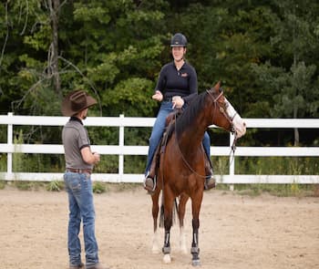 A person on horseback is receiving instructions from another person standing nearby in an outdoor riding arena. The rider is wearing a black shirt and helmet, while the instructor is wearing a cowboy hat and casual attire. There is a white fence and green foliage in the background.