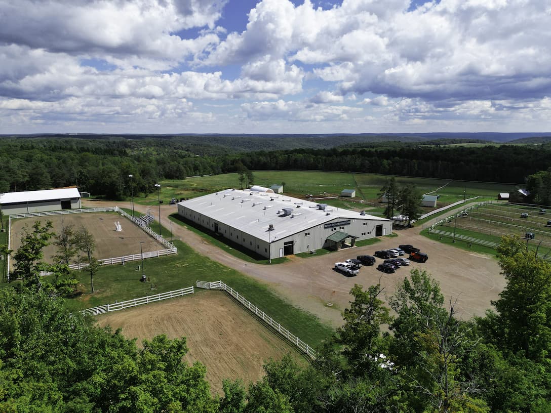 Aerial view of a modern equestrian center with a large white-roofed stable, outdoor riding arena, and surrounding green pastures and forest. Located in a scenic rural area, the horse facility includes multiple fenced paddocks, support buildings, and a gravel parking area with vehicles. Ideal for horseback riding, training, and equine events.