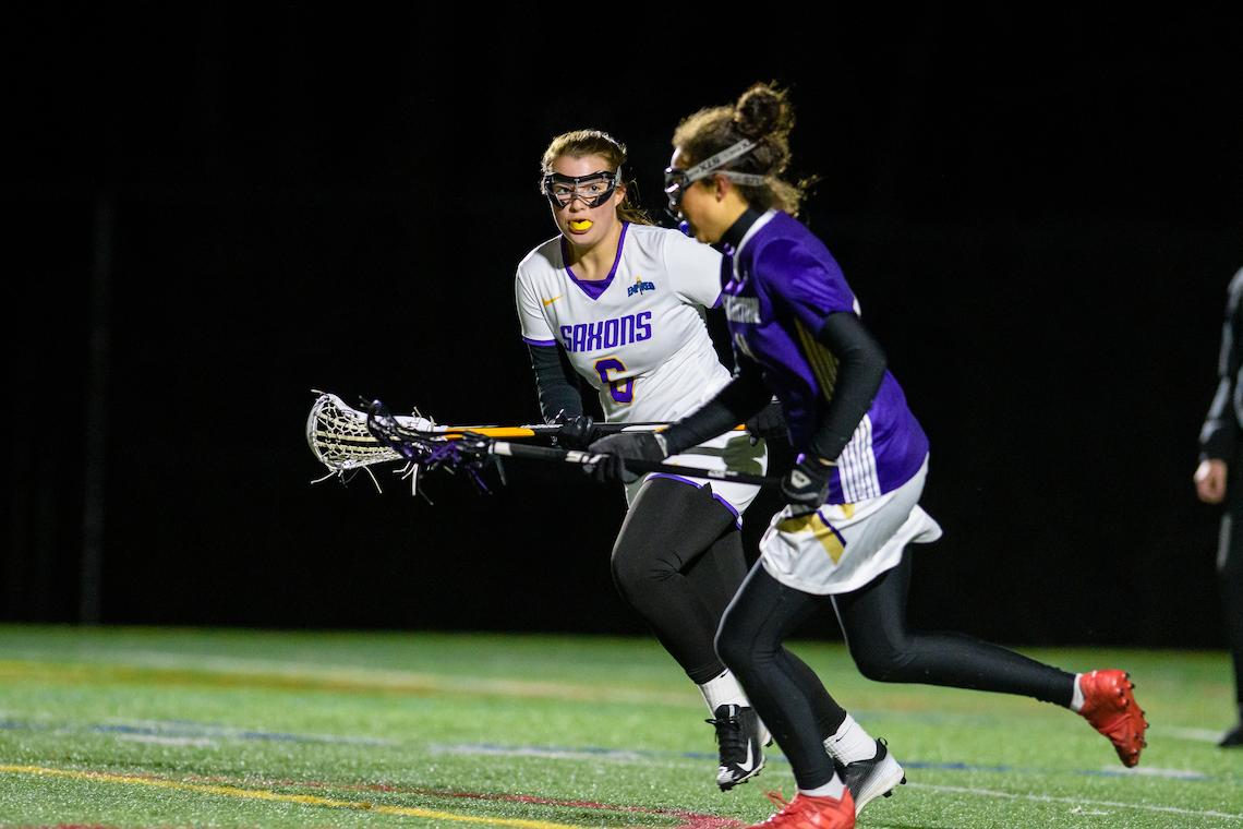 Two lacrosse players compete on a field at night. One, in a white 'Saxons' jersey with number 6, holds a lacrosse stick, while the other, in a purple jersey, runs with the ball. Both wear black leggings and cleats, with one pair of cleats in red.