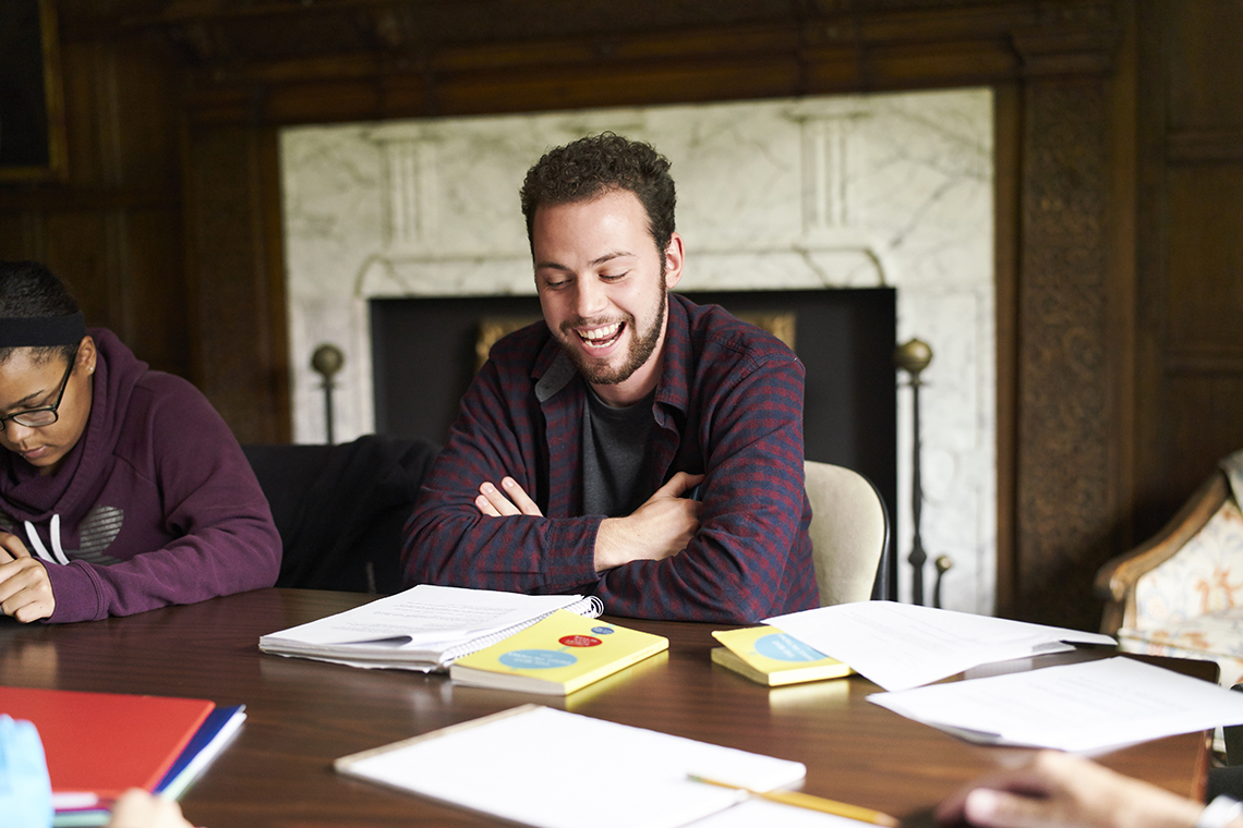 A person in a dark T-shirt focuses on operating a milling machine in a workshop. Soft light from a window casts shadows, creating an industrious mood.
