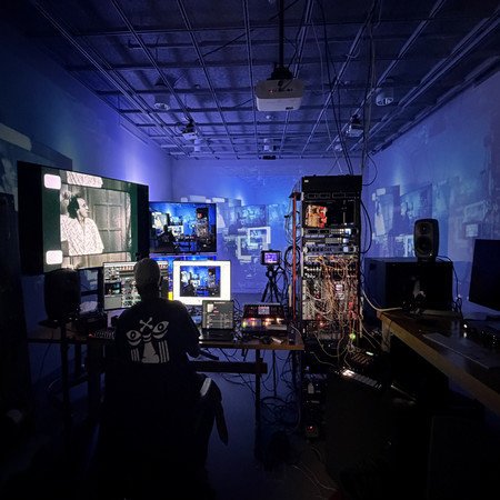 A dark room with immersive projection art on walls, depicting hands and abstract organic shapes. A person stands surrounded by circular lights, creating a mysterious, contemplative atmosphere. vy Ali Santana