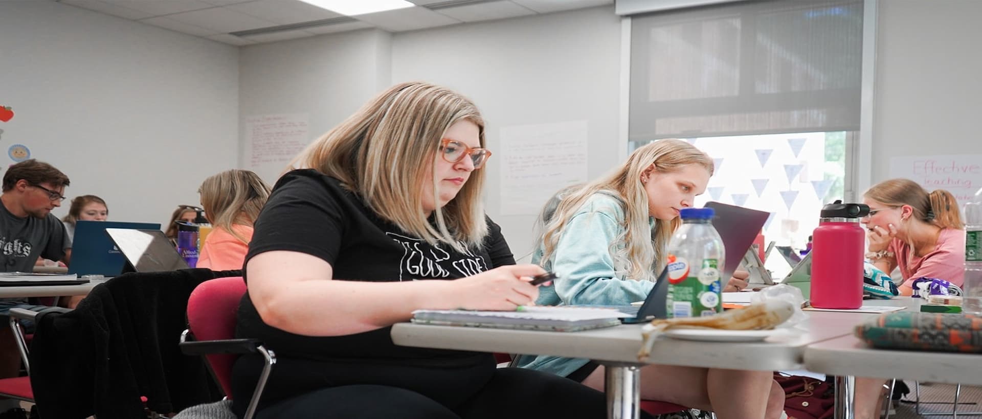 Students sitting at desks in a classroom, working on assignments with notebooks, laptops, and water bottles on the tables.