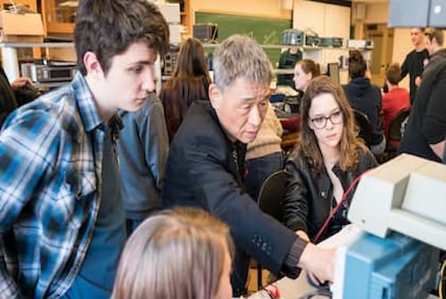 A group of people gathers around electronic equipment in a classroom or lab setting. Some are seated while others stand, all focused on the device. Shelves with various devices and a chalkboard are visible in the background.