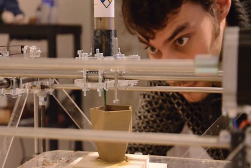 A person closely watches a 3D printer creating a small, angular object with brown material. The background includes blurred shelves and equipment in a workspace.