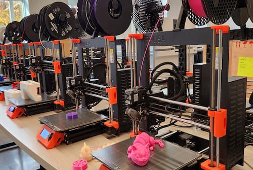 A row of 3D printers on a table, each with filament spools above them, actively printing objects. A pink model is visible in the foreground, and a purple object is being printed on another machine. The background includes equipment and supplies in a well-lit room.