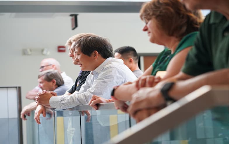 A group of casually dressed people leans on a glass railing indoors, looking downward. Behind them is a wall with fixtures and a partially visible door.