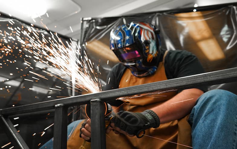 A person wearing a flame-patterned welding helmet, gloves, and a protective apron is using a power tool to grind metal, creating a shower of sparks. They are working on a black metal structure in a workshop with dark protective curtains in the background, highlighting a hands-on fabrication or welding environment.