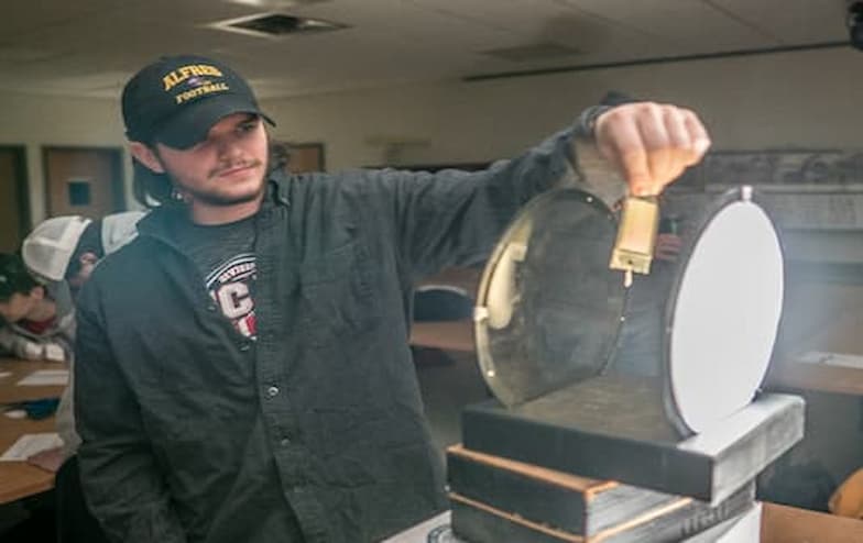 A person wearing a dark shirt and a cap labeled "ALFRED" holds a small cylindrical object in front of a large circular mirror or lens. They appear to be demonstrating an optics experiment in a classroom or lab setting. In the background, others are seated at tables with books and equipment, observing the demonstration.