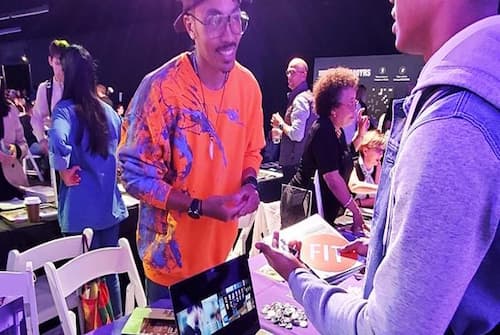 People are interacting at a busy indoor event with tables displaying various items. One person in an orange and blue shirt stands next to a table with a laptop showing images, while another person in a gray hoodie holds a booklet. Other attendees are visible in the background, engaging in conversations.