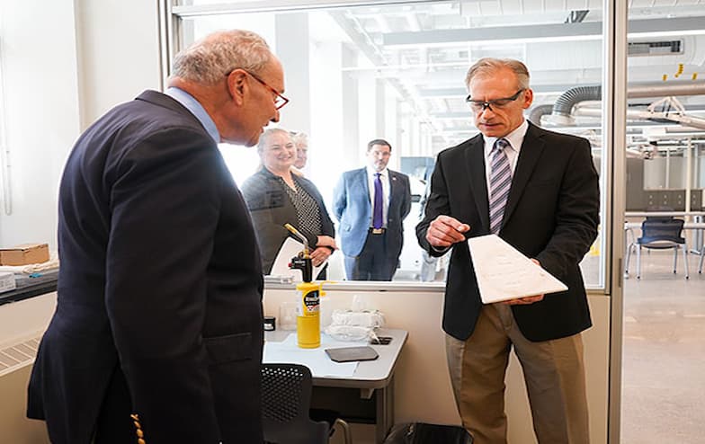A man in a black suit and striped tie holds and explains a white, triangular research prototype to another man in a blue suit and glasses. They are inside a modern lab space with glass walls, while three professionally dressed individuals observe from outside the room. A can of Lysol wipes and lab equipment are visible on a nearby desk, suggesting a clean and controlled research environment.