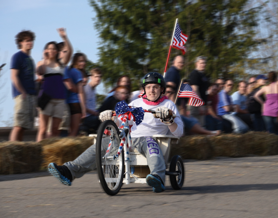 Student racing a decorated cart in the Pine Hill Derby while spectators cheer from the sidelines.