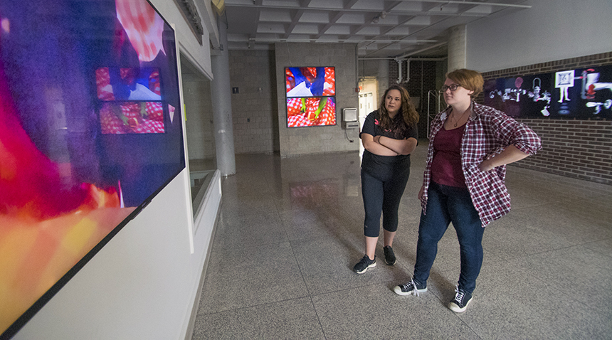 Two people stand in a hallway gallery space viewing large, colorful digital artworks displayed on wall-mounted screens.