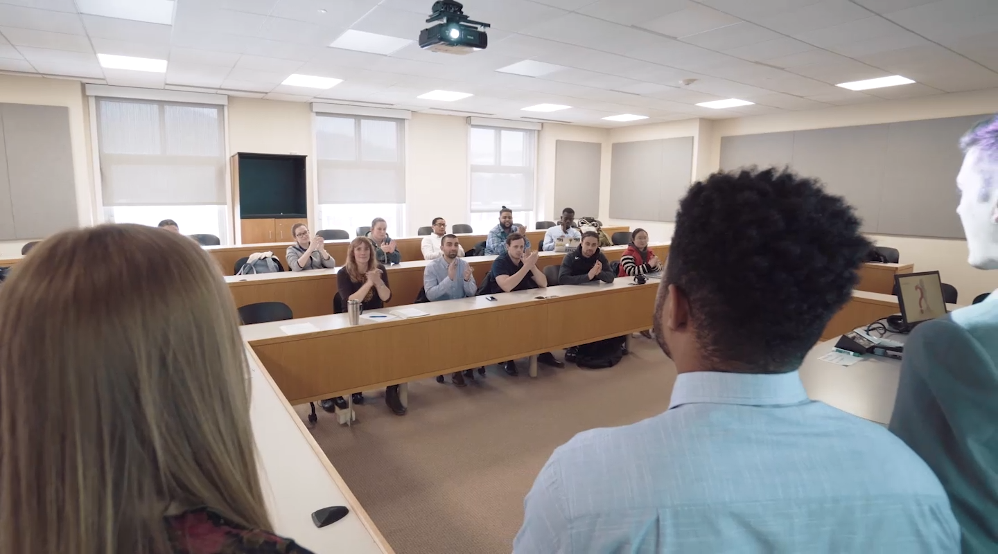 Students in a business classroom applaud presenters standing at the front of the room.