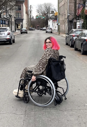 woman with bright pink hair looking over her shoulder toward the camera, sitting in a wheel chair in the middle of a city street