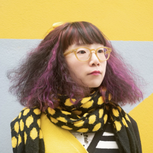 woman with pruple streaked sholder length hair wearing black and yellow polka dot scarf pictured on a background of yellow and gray